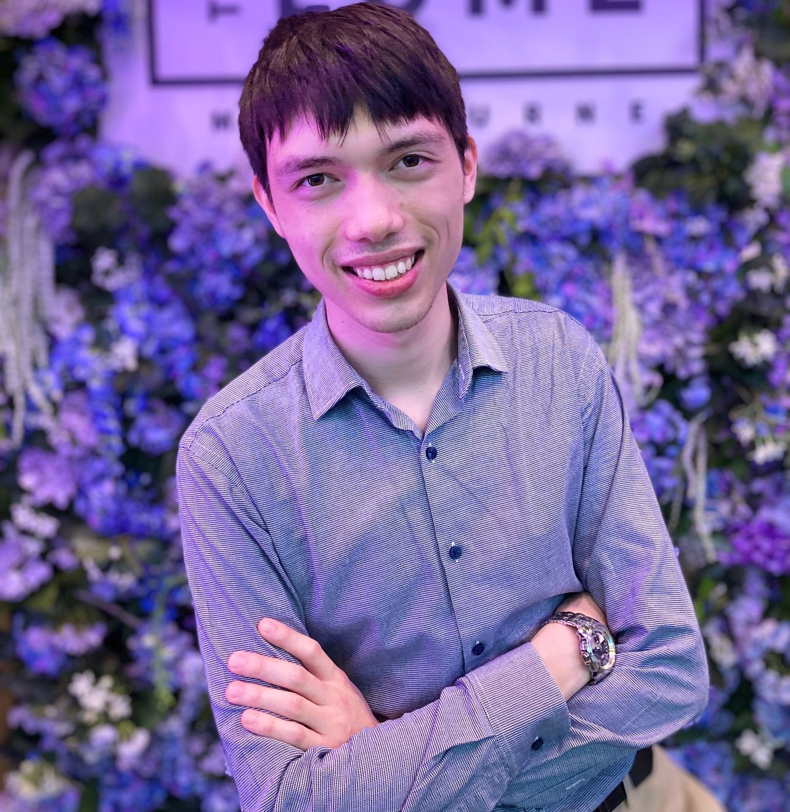 Young man stands with arms folded in front of a wall of blue and purple flowers