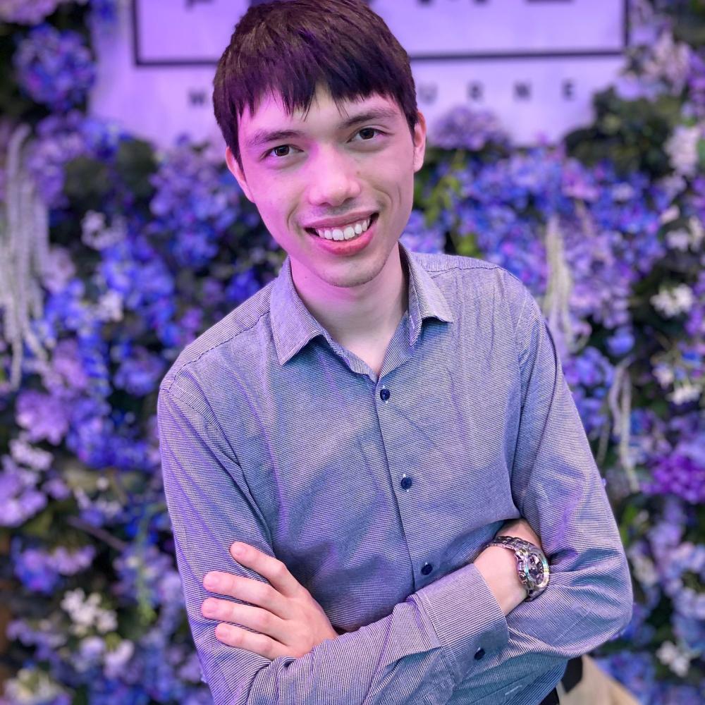 Young man stands with arms folded in front of a wall of blue and purple flowers